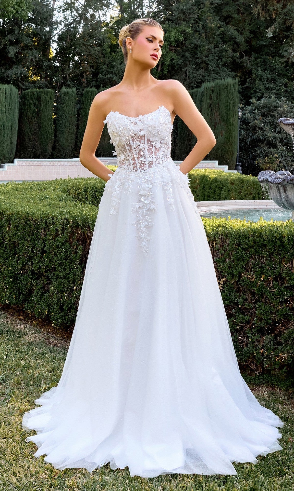 A woman in the Ladivine C174W off white formal dress, featuring a strapless corset bodice and delicate 3D floral appliqué, stands outdoors in a manicured garden.